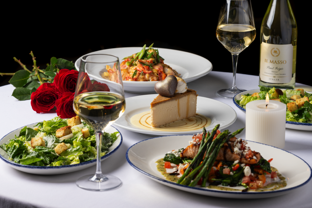 Elegant Valentine's Day dinner featuring salmon with asparagus, Caesar salad, pasta, cheesecake, wine, and red roses on a white tablecloth.